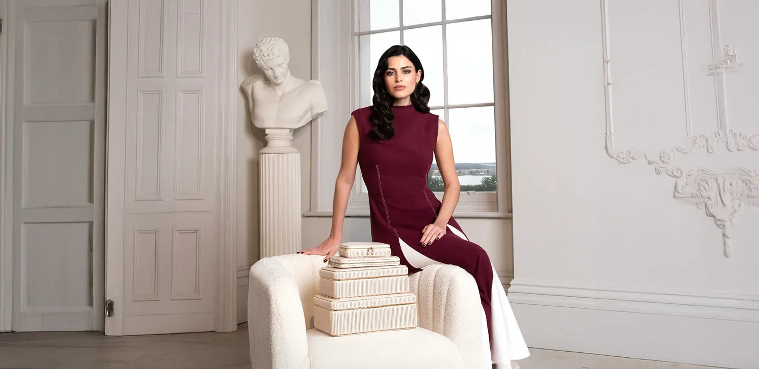 Eine elegant gekleidete Frau in einem burgunderroten Kleid sitzt neben einer Auswahl an luxuriösen, cremefarbenen Schmuckschatullen der WOLF Bella Kollektion mit plissierter Oberfläche, präsentiert in einem stilvollen Raum mit klassischer Büste – erhältlich im juwelier-tirol.shop.