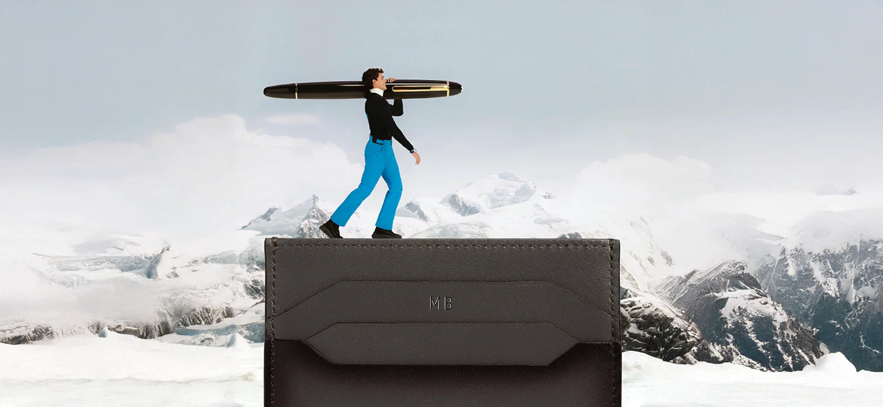 Eine surrealistische Montage zeigt eine kleine männliche Figur in blauen Hosen, die ein riesiges Montblanc Meisterstück Schreibgerät über eine verschneite Gebirgslandschaft trägt. Die Szene spielt sich auf der Oberkante einer braunen Ledergeldbörse ab. Erhältlich im juwelier-tirol.shop.