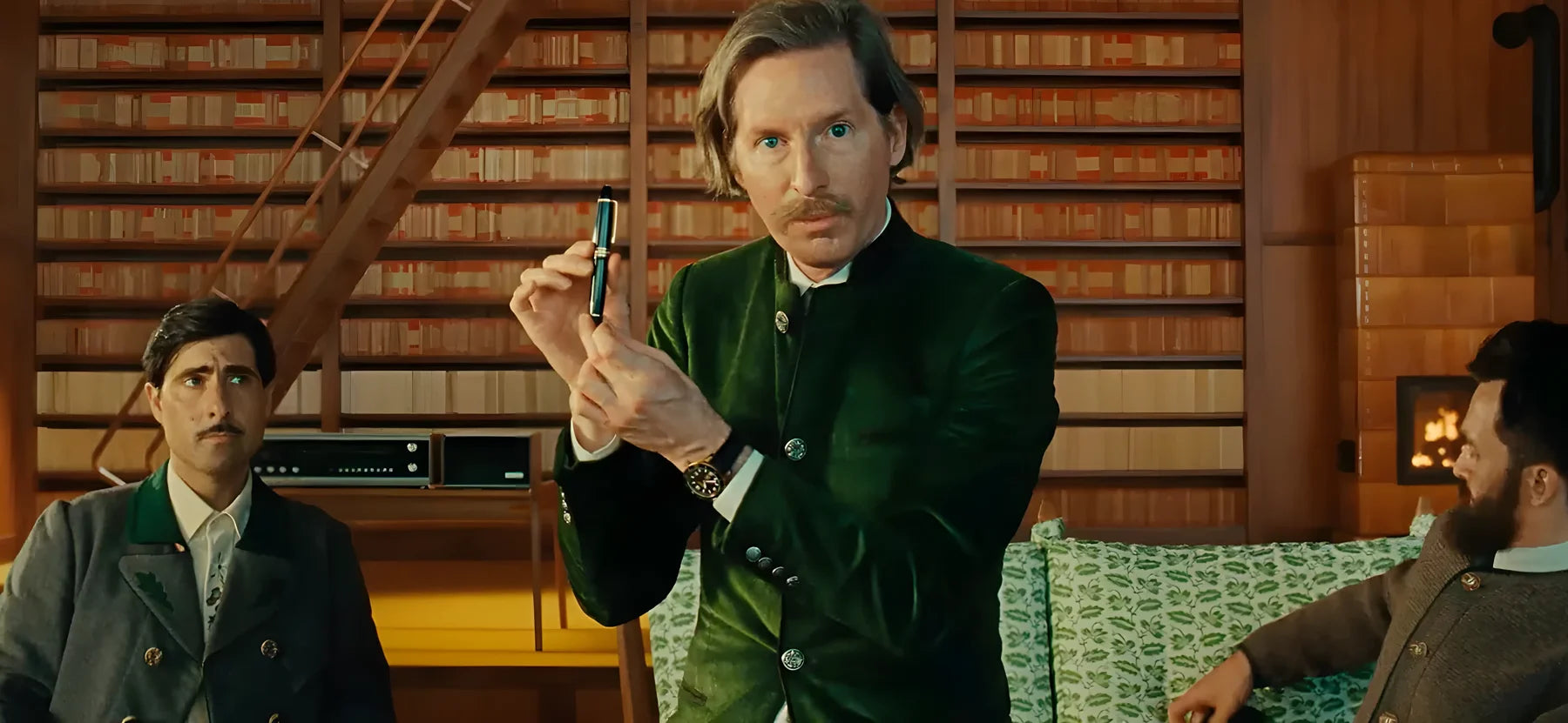 Der Regisseur Wes Anderson im dunkelgrünen Samtsakko präsentiert einen edlen Schreibstift. Im Hintergrund sitzen zwei Männer vor einer riesigen Bücherwand in einem warm beleuchteten Raum. Erhältlich im juwelier-tirol.shop